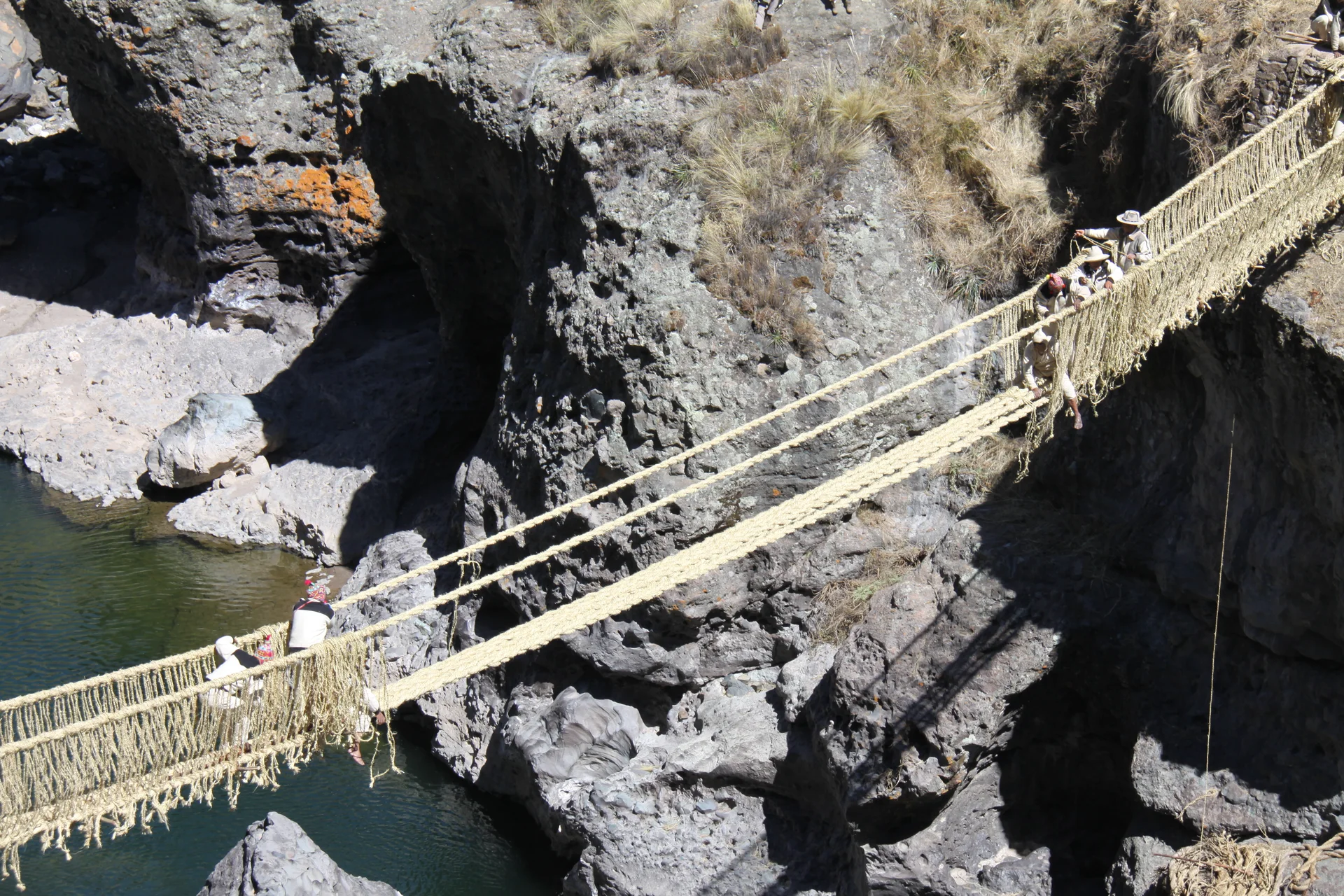Bridge builders work on the woven Q’eswachaka bridge above the water between steep rock walls.